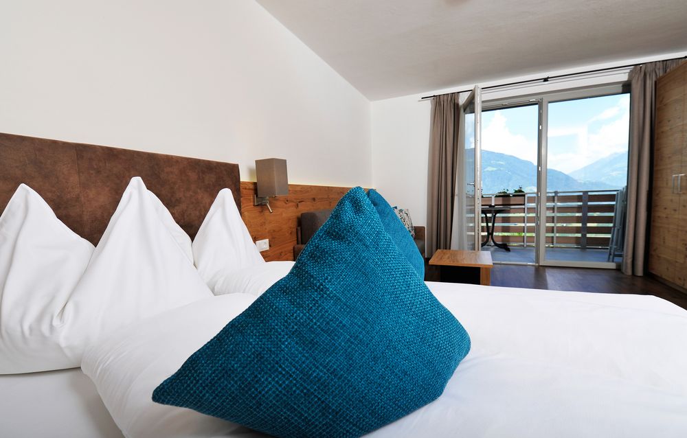 Modernes Hotelzimmer mit blauem Kissen, weißer Bettwäsche und Balkon mit Bergblick.