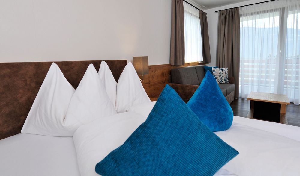 Modernes Schlafzimmer mit braunem Bett, weißen und blauen Kissen, Sofa und großen Fenstern mit Vorhängen.