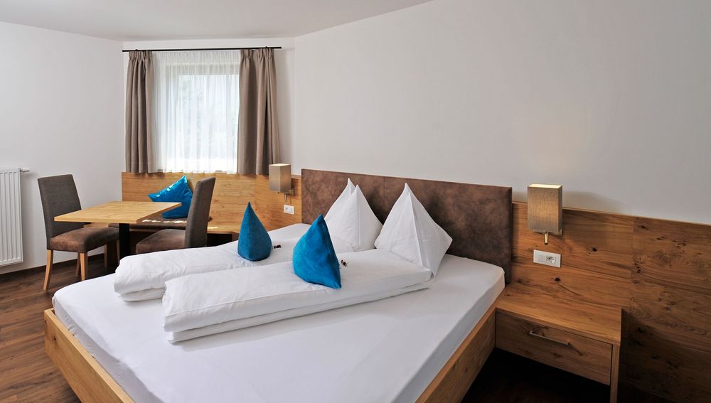 Gemütliches Hotelzimmer mit Doppelbett, blauen Kissen und Sitzecke am Fenster