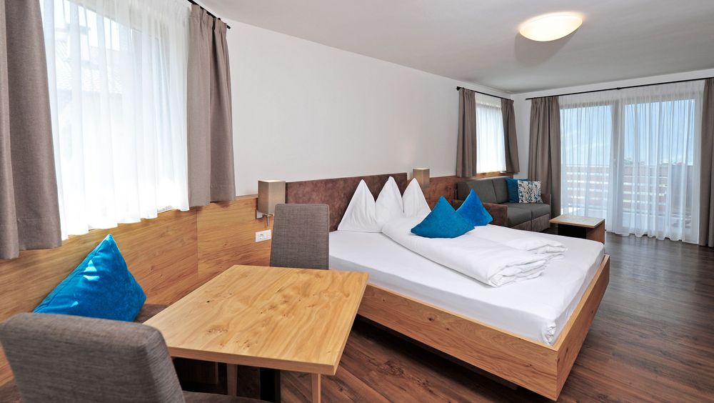 Modernes Schlafzimmer mit Holz- und weißen Wänden, Bett, Tisch und Stühlen, Sofa und großem Fenster mit Vorhängen
